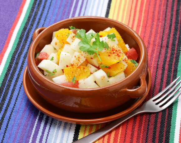 Terra cotta colored bowl filled with a salad composed of oranges and jicama placed on a colorful striped background.