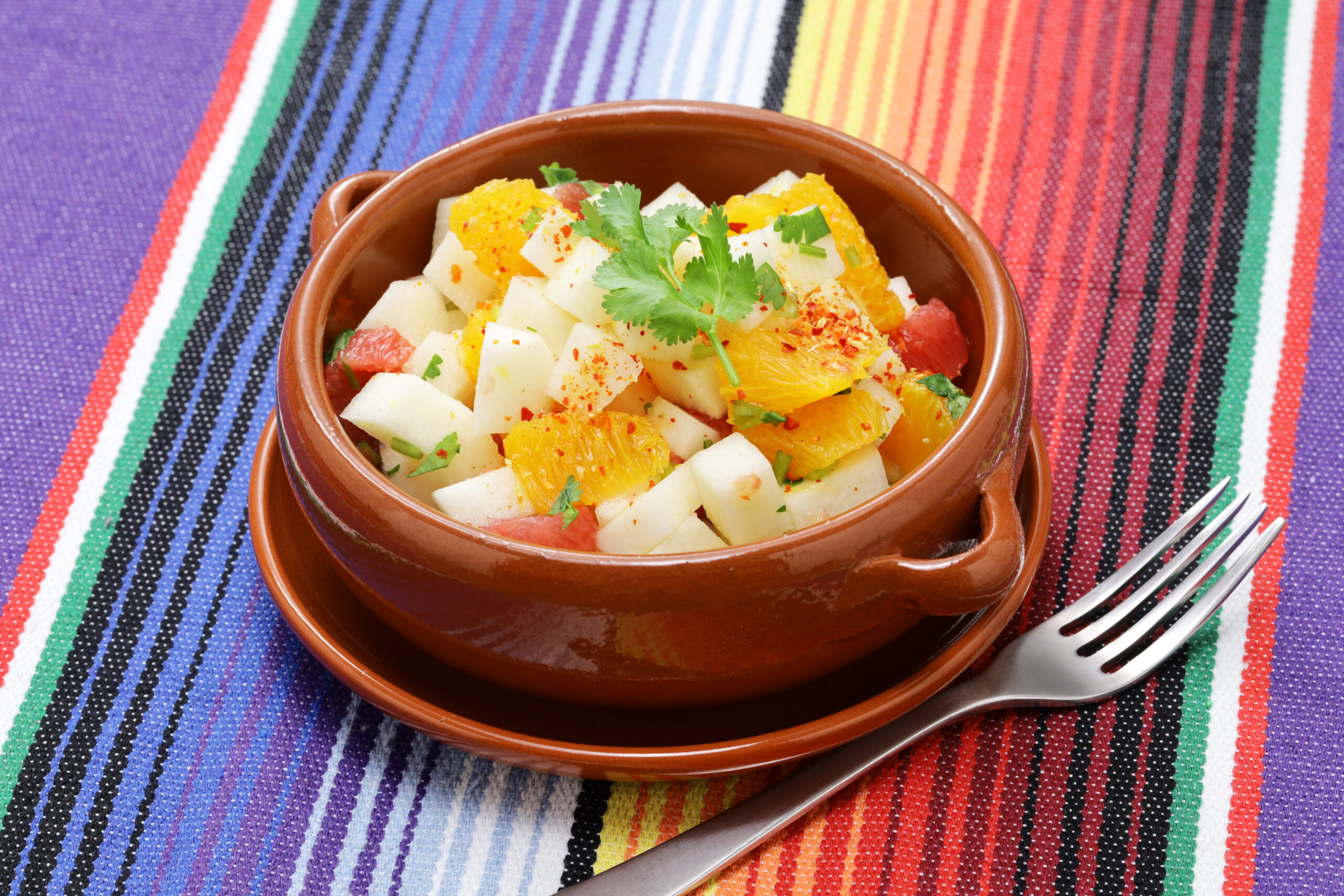 Terra cotta colored bowl filled with a salad composed of oranges and jicama placed on a colorful striped background.